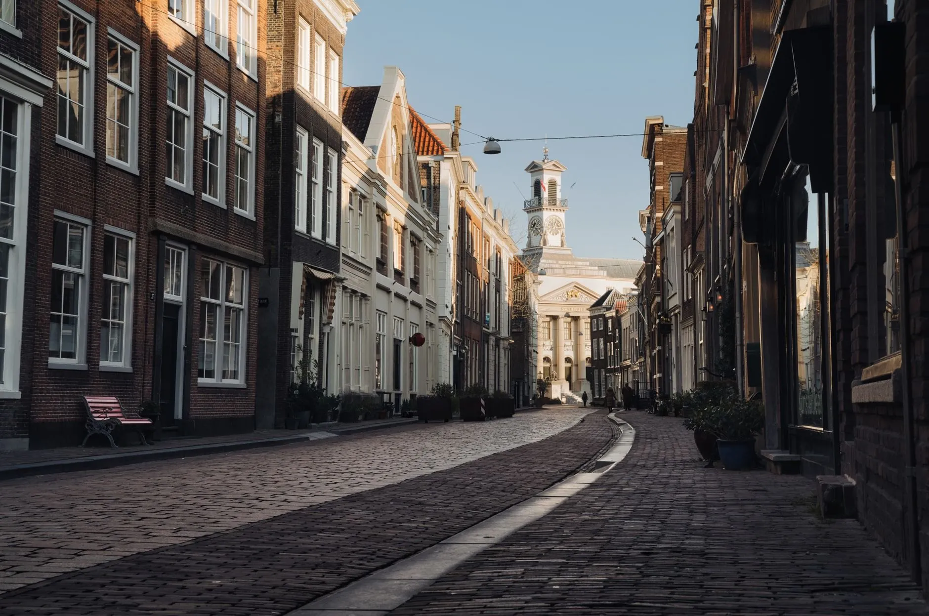 An empty cobbled street of tall brick houses leading to a white clock tower in early sun