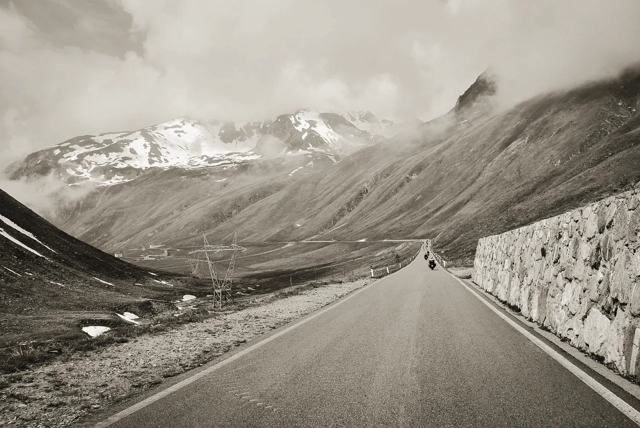 A mountain pass road curving under snow-streaked alpine peaks and cloud